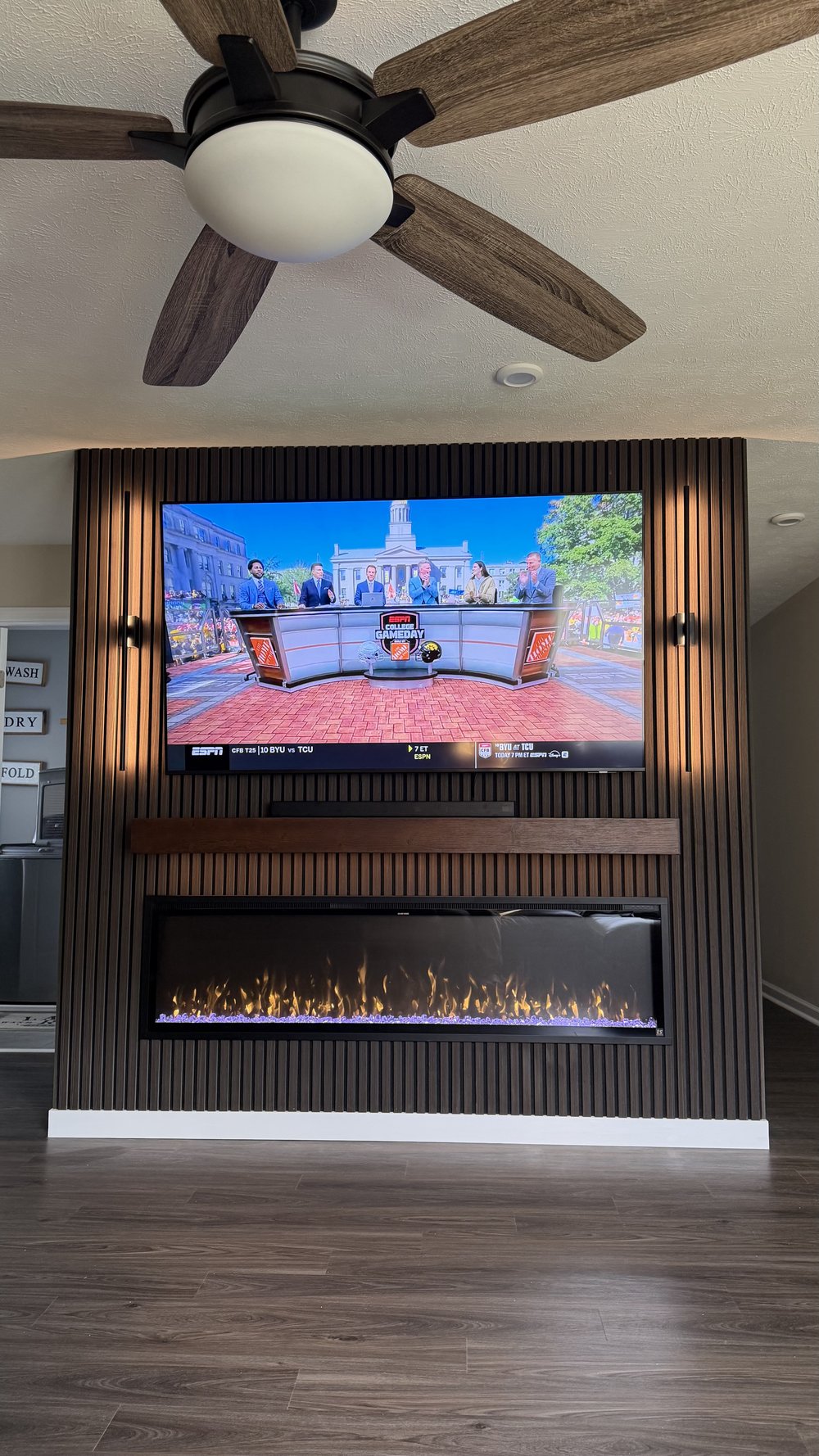 Dark wood slat media wall with mounted TV, floating mantel, fireplace, and black wall sconces